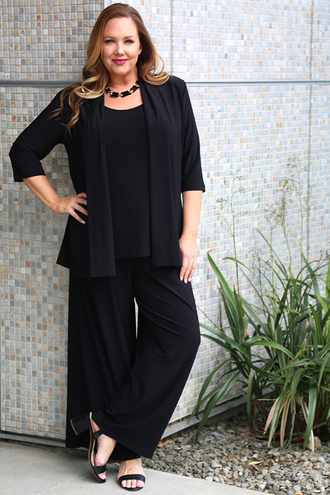 Woman in a black outfit standing against a tiled wall with plants.