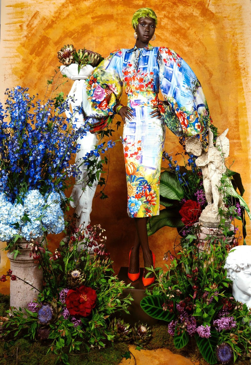 Person in a colorful outfit surrounded by flowers and statues against an orange background