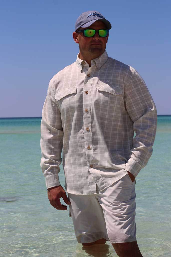 Man standing in clear water wearing a light shirt, shorts, and cap with sunglasses on a sunny day.