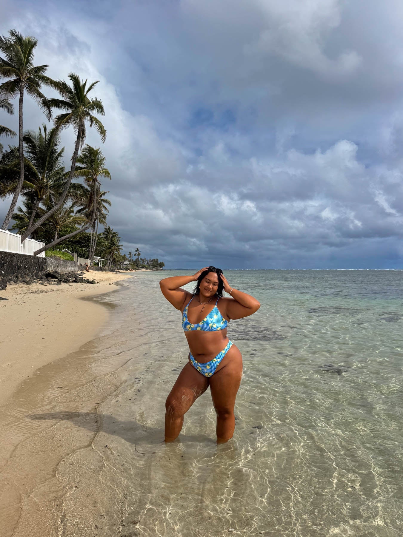 Woman in a blue bikini standing on a beach with palm trees and clear water.