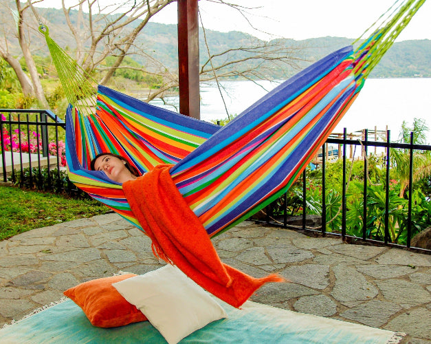 Person relaxing in a colorful striped hammock by a body of water.