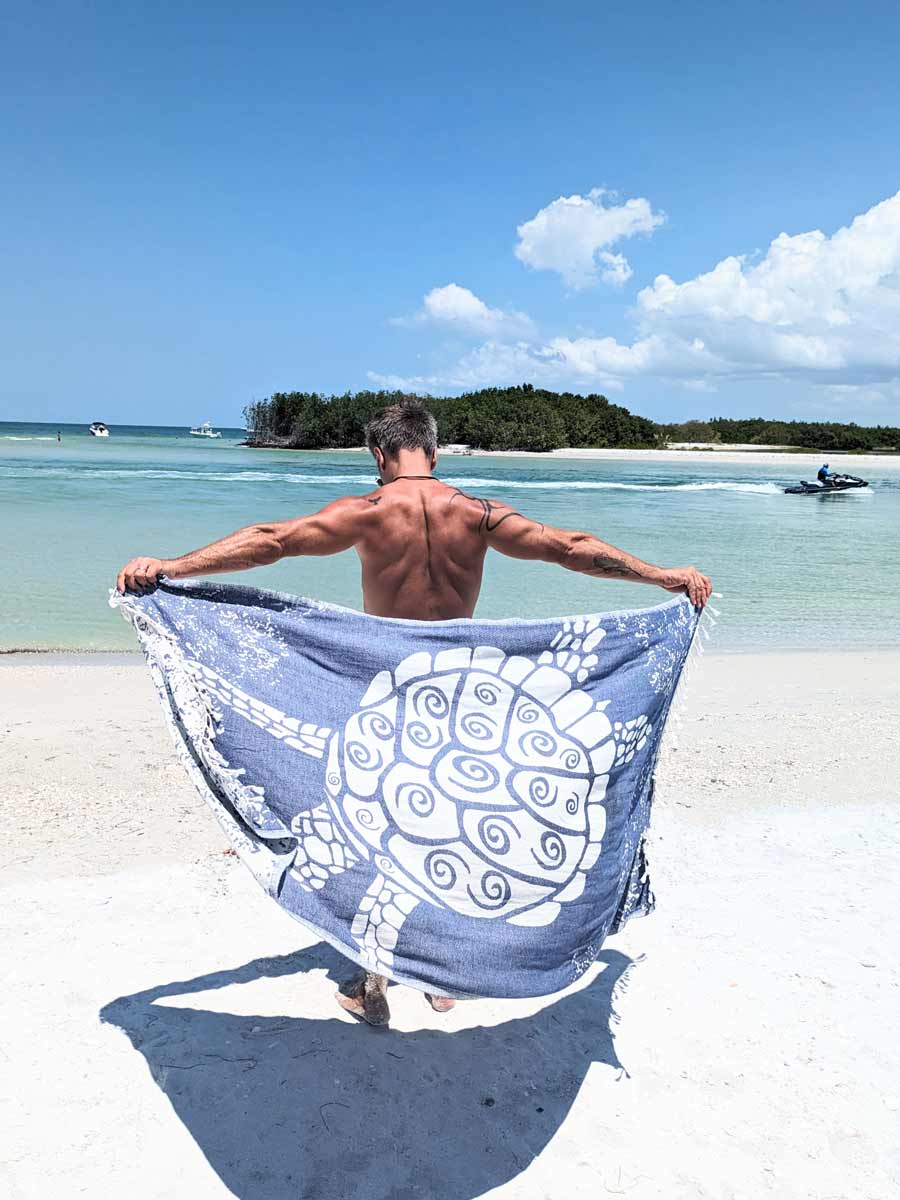 Man holding a blue and white patterned towel on a beach with clear blue water and sky.