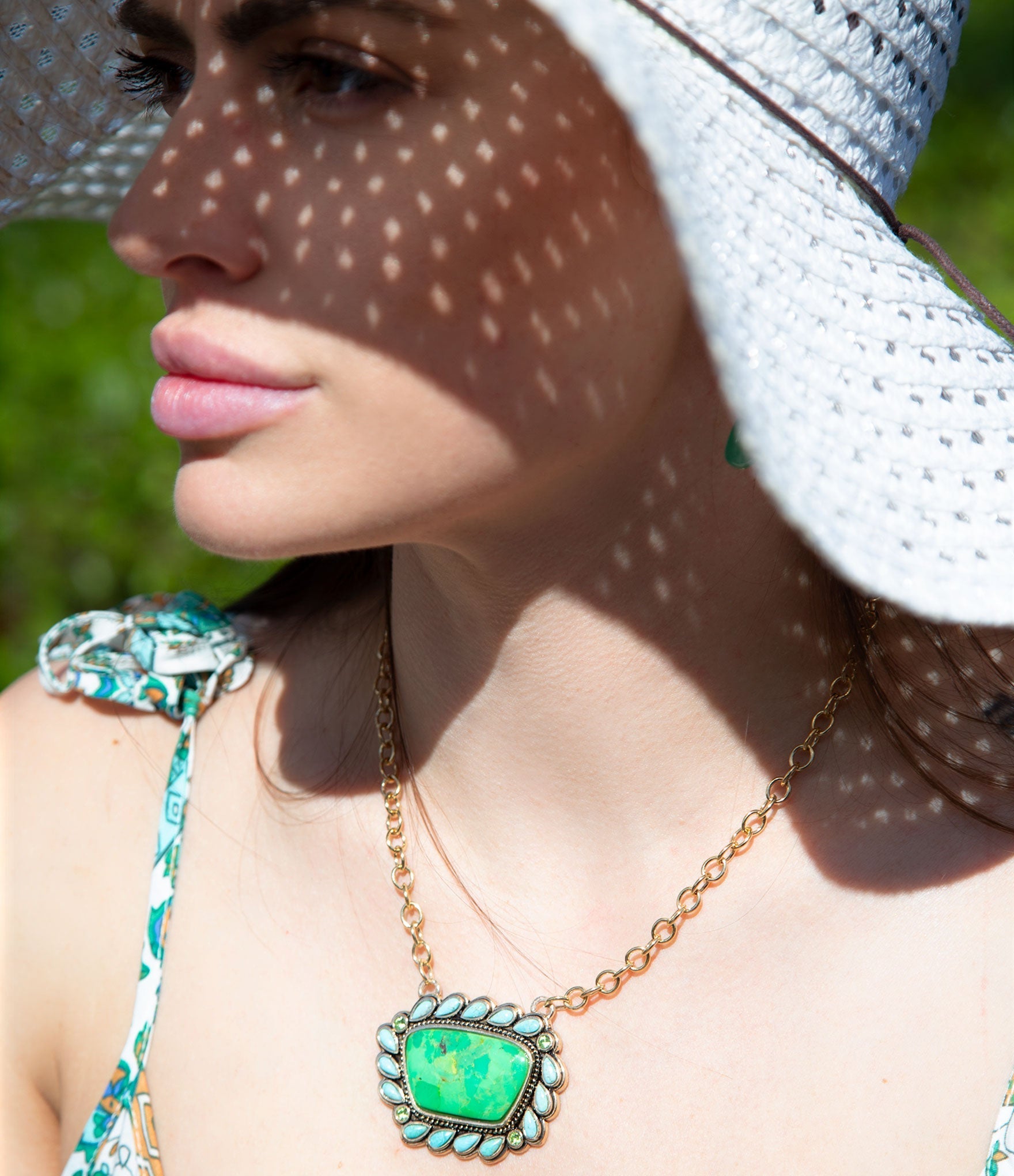 Woman wearing a white sun hat and green necklace with a large pendant, standing outdoors.