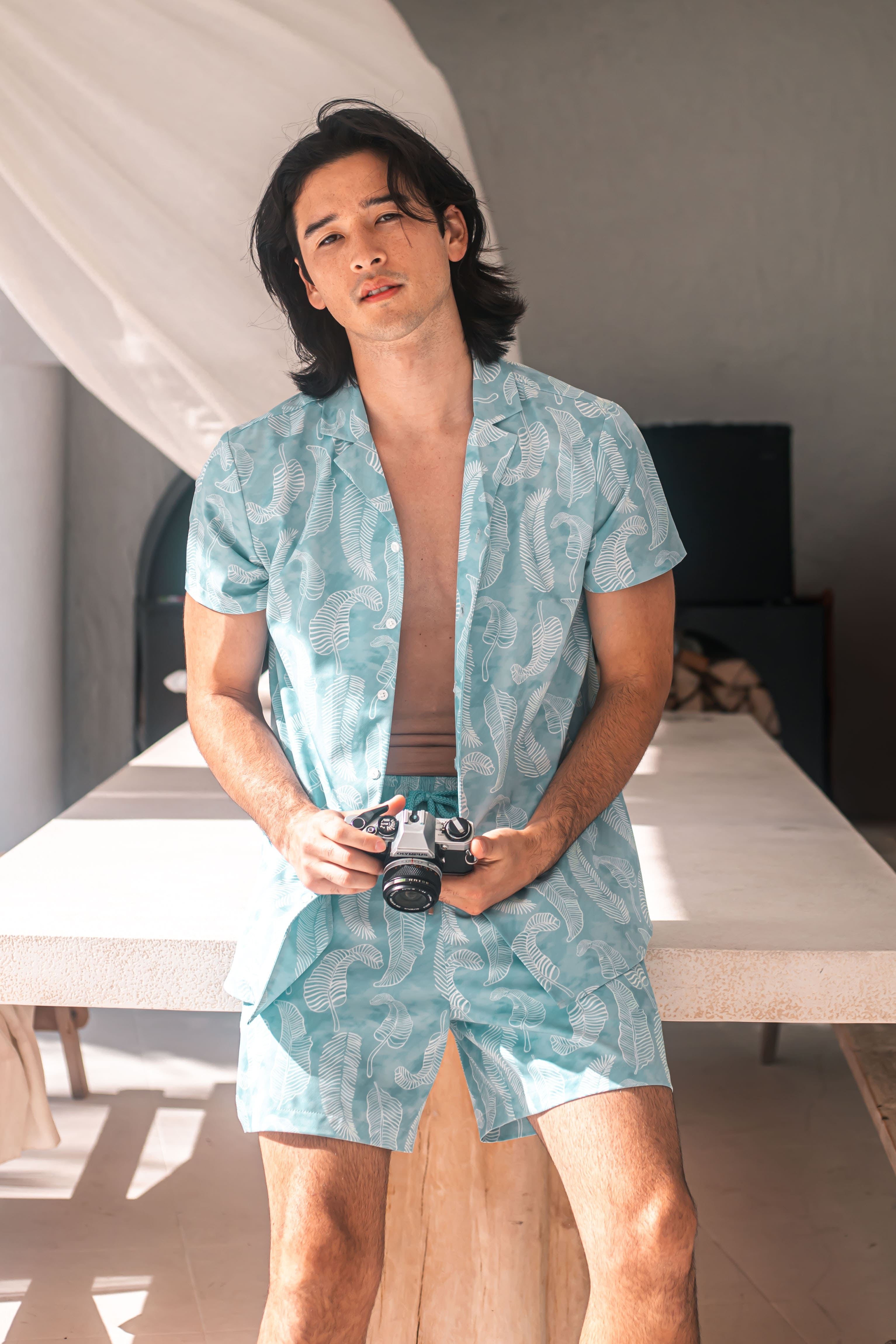 Man wearing a light blue patterned shirt and shorts holding a camera indoors.