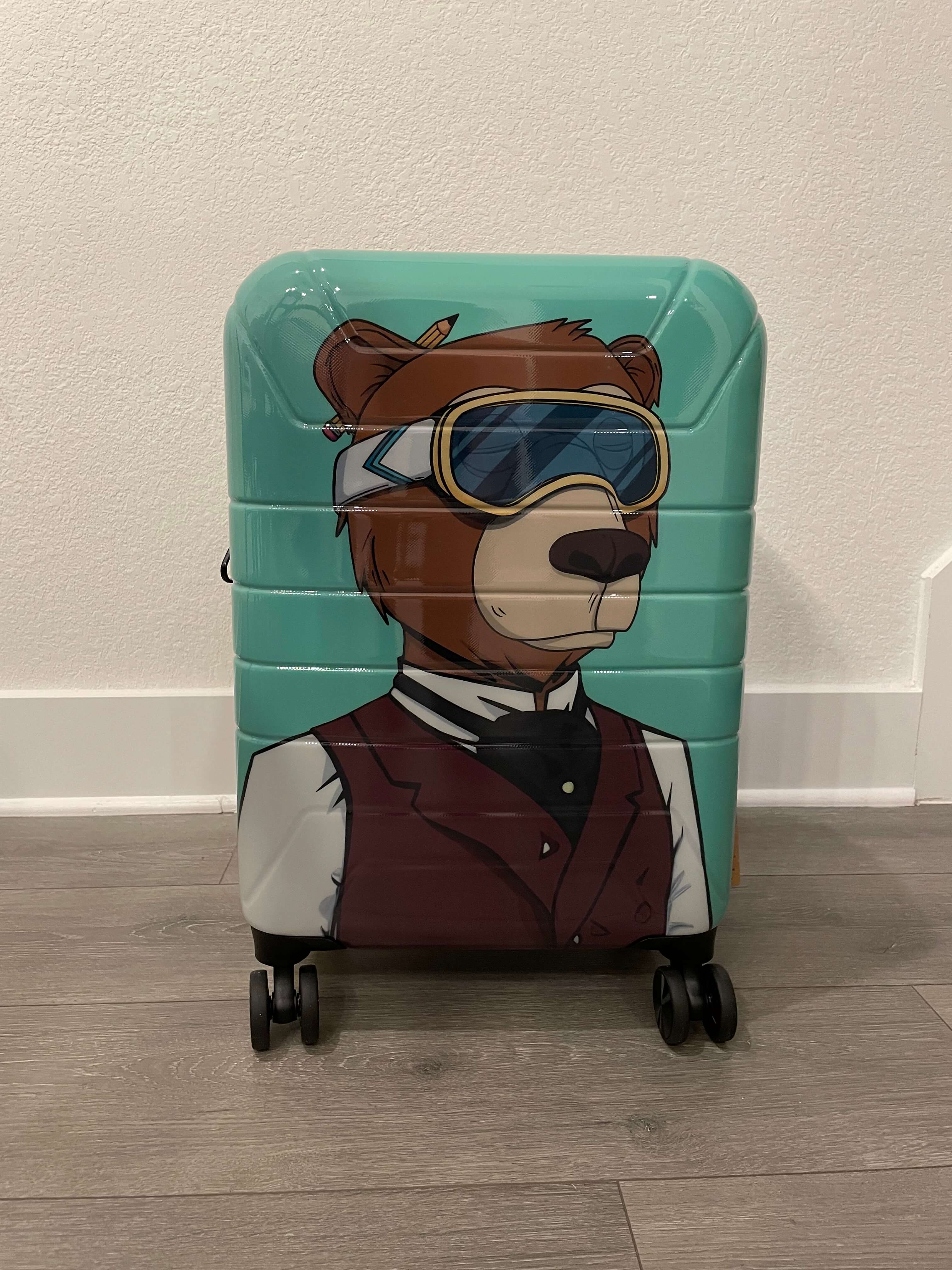Suitcase with a bear design wearing goggles and a suit on a wooden floor.