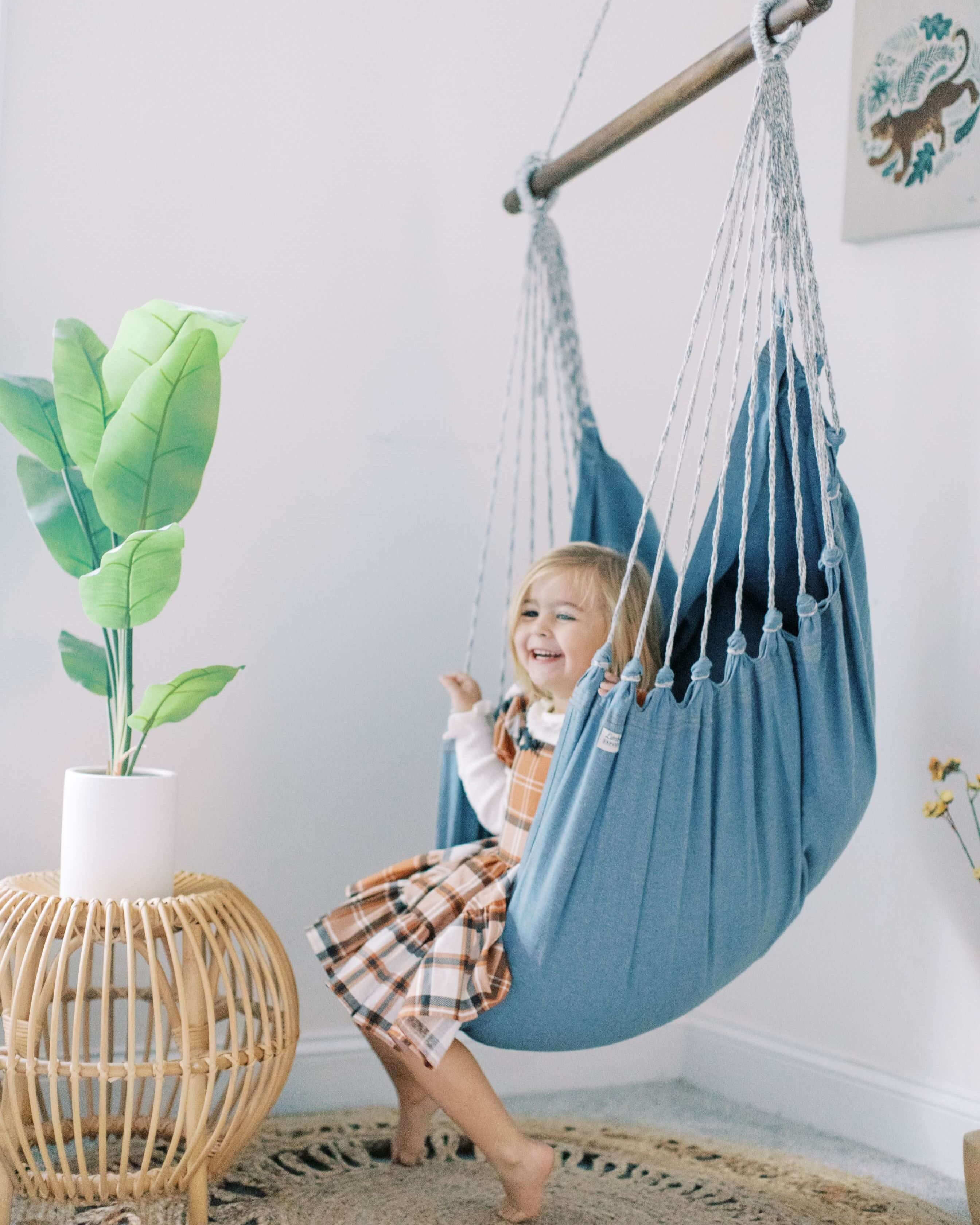 Child in a blue hammock chair in a room with a plant and table.