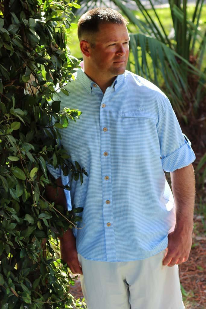 Man wearing a light blue shirt and white pants standing next to green foliage