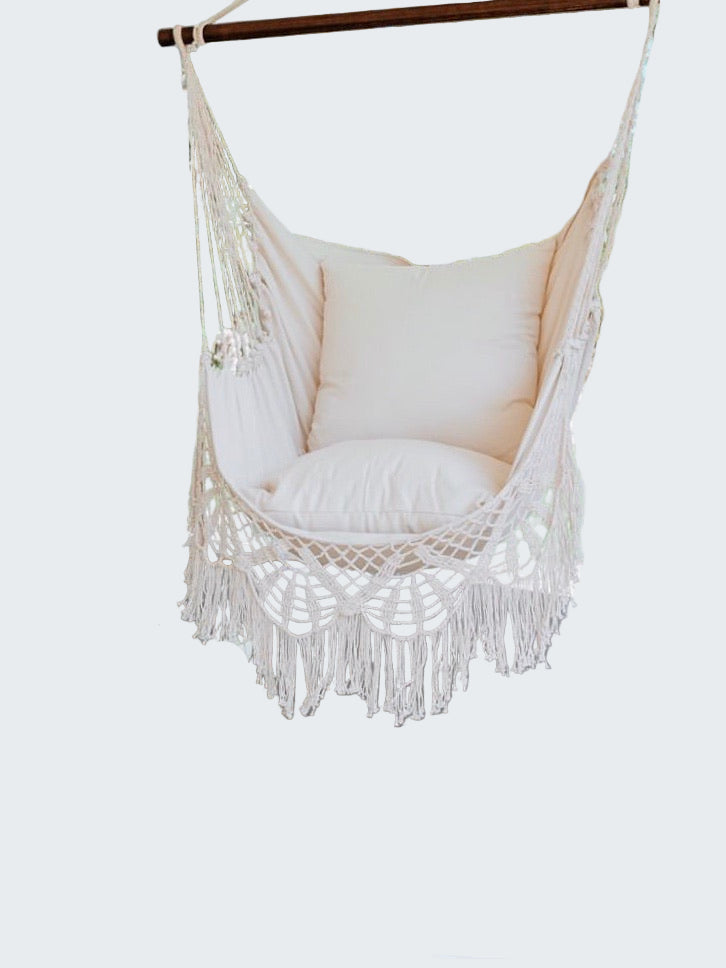 Macrame hammock chair with cushions on a wooden deck, surrounded by greenery.