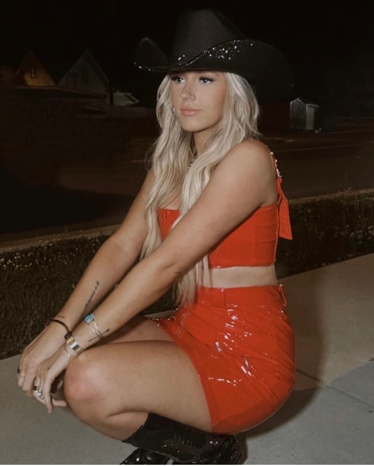 Woman in a red outfit and black cowboy hat sitting on a curb at night.