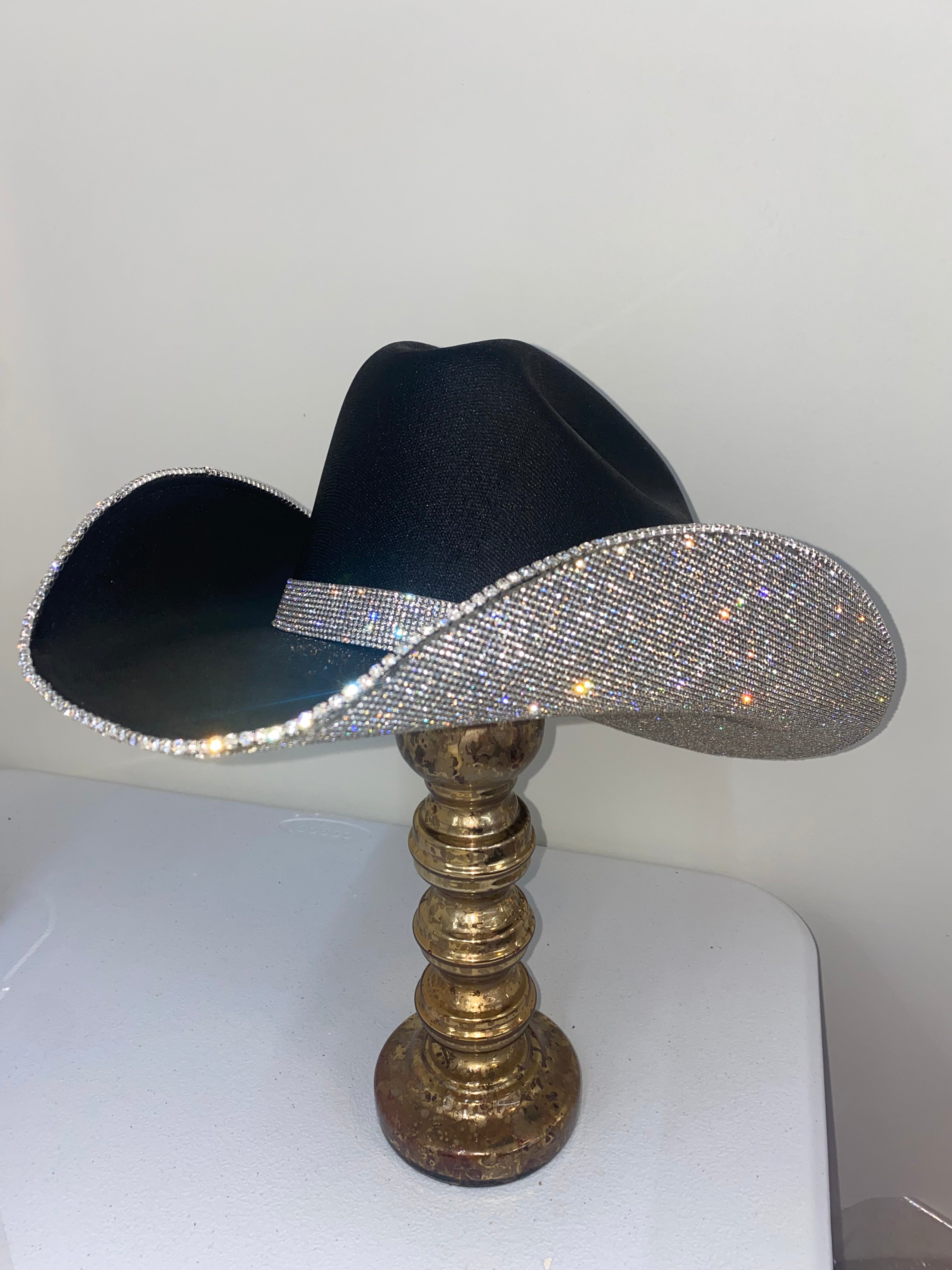 Black cowboy hat with a rhinestone band on a gold stand against a white background