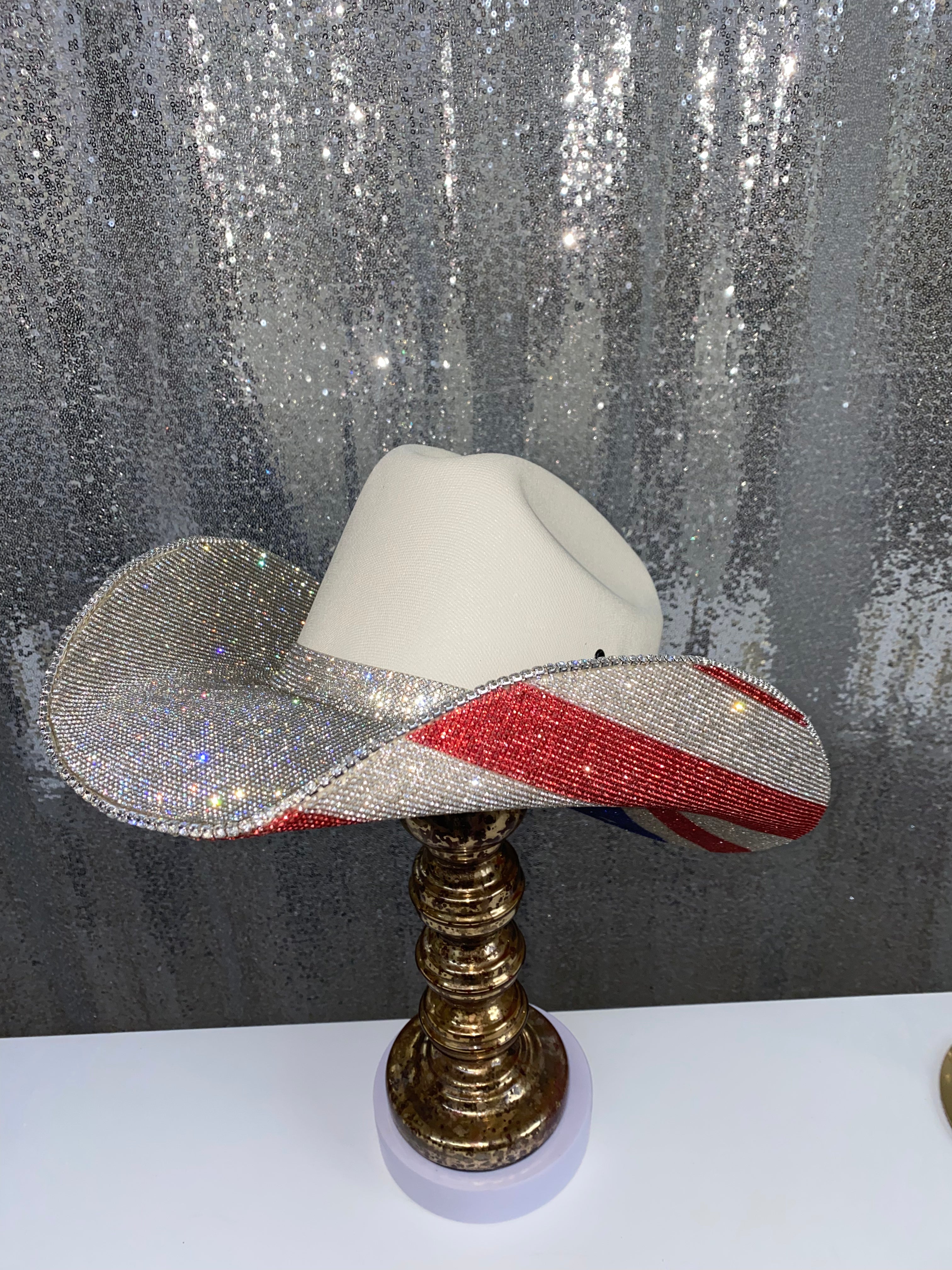 Decorative cowboy hat with red, white, and blue design on a stand against a glittery curtain background.