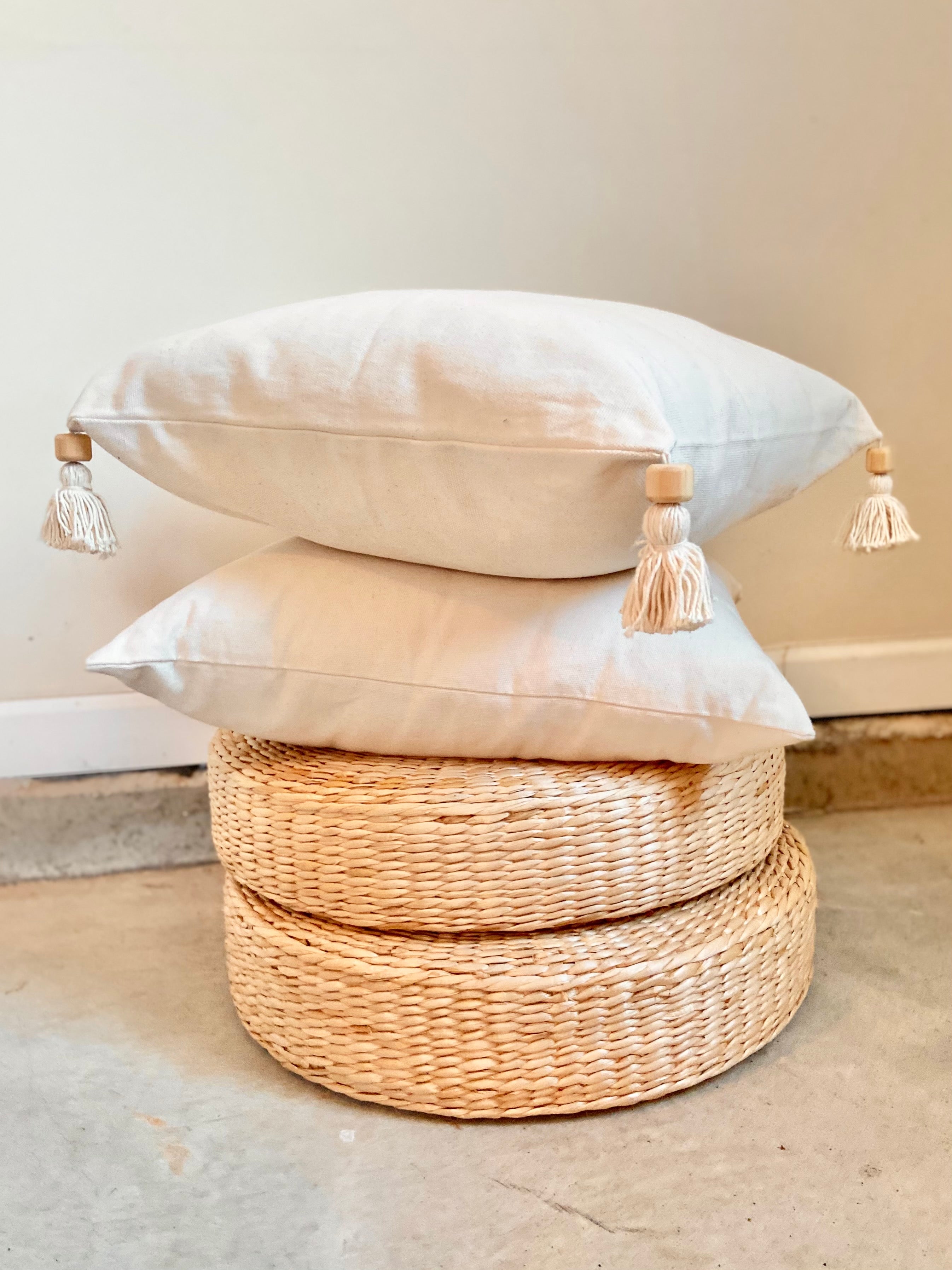 Stack of decorative pillows with tassels on a woven stool against a neutral wall.