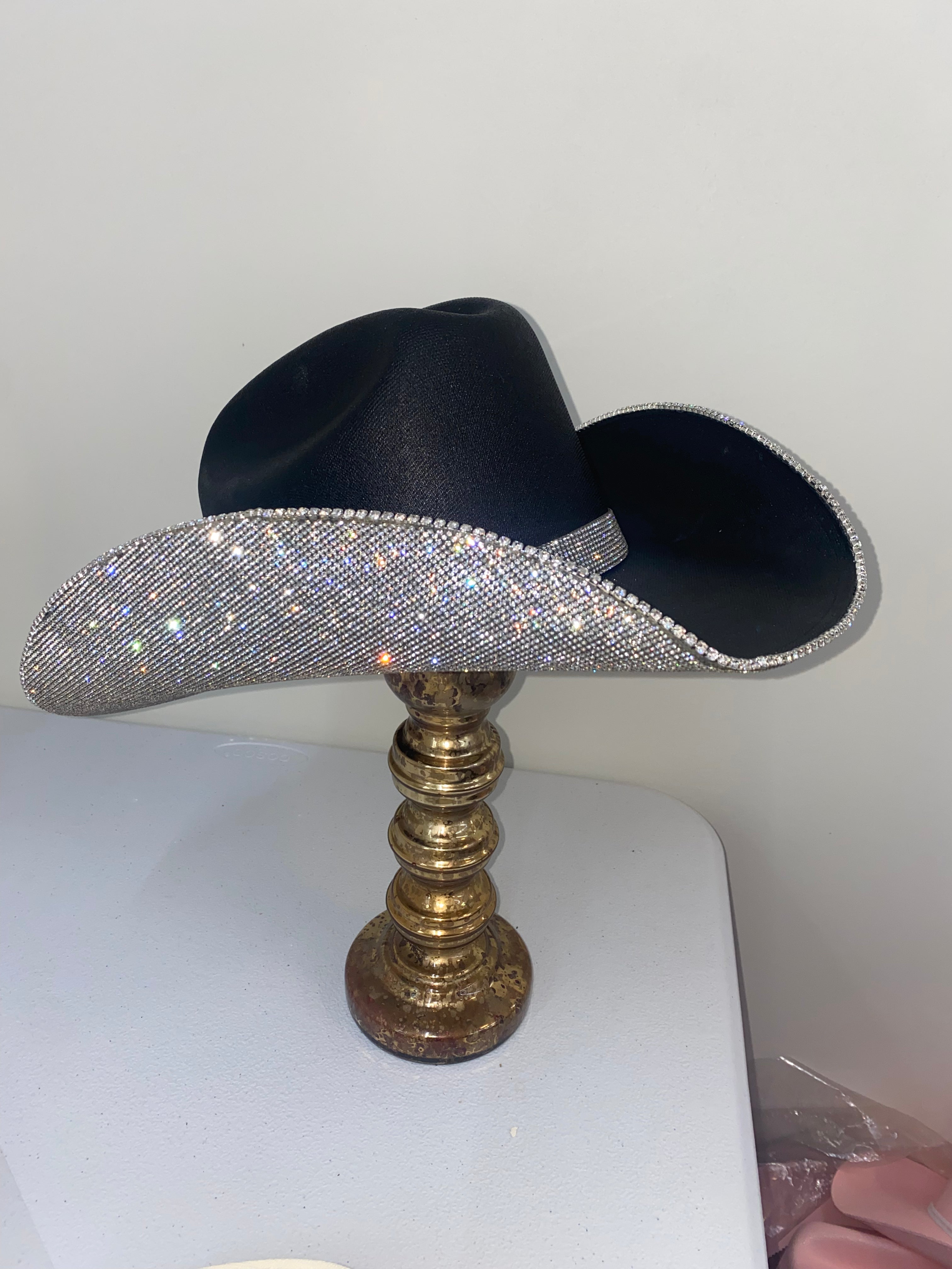 Black cowboy hat with a rhinestone band on a gold stand against a white background