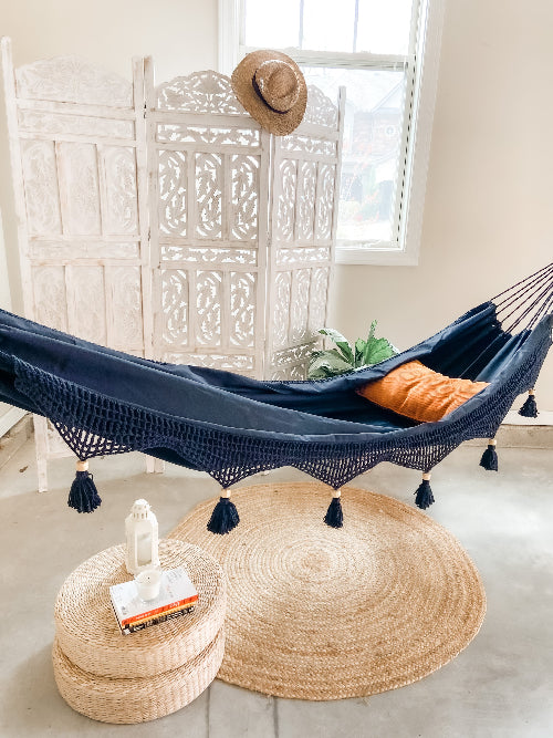 Navy blue hammock with orange pillow on a woven mat in a room with a white lattice screen and window.