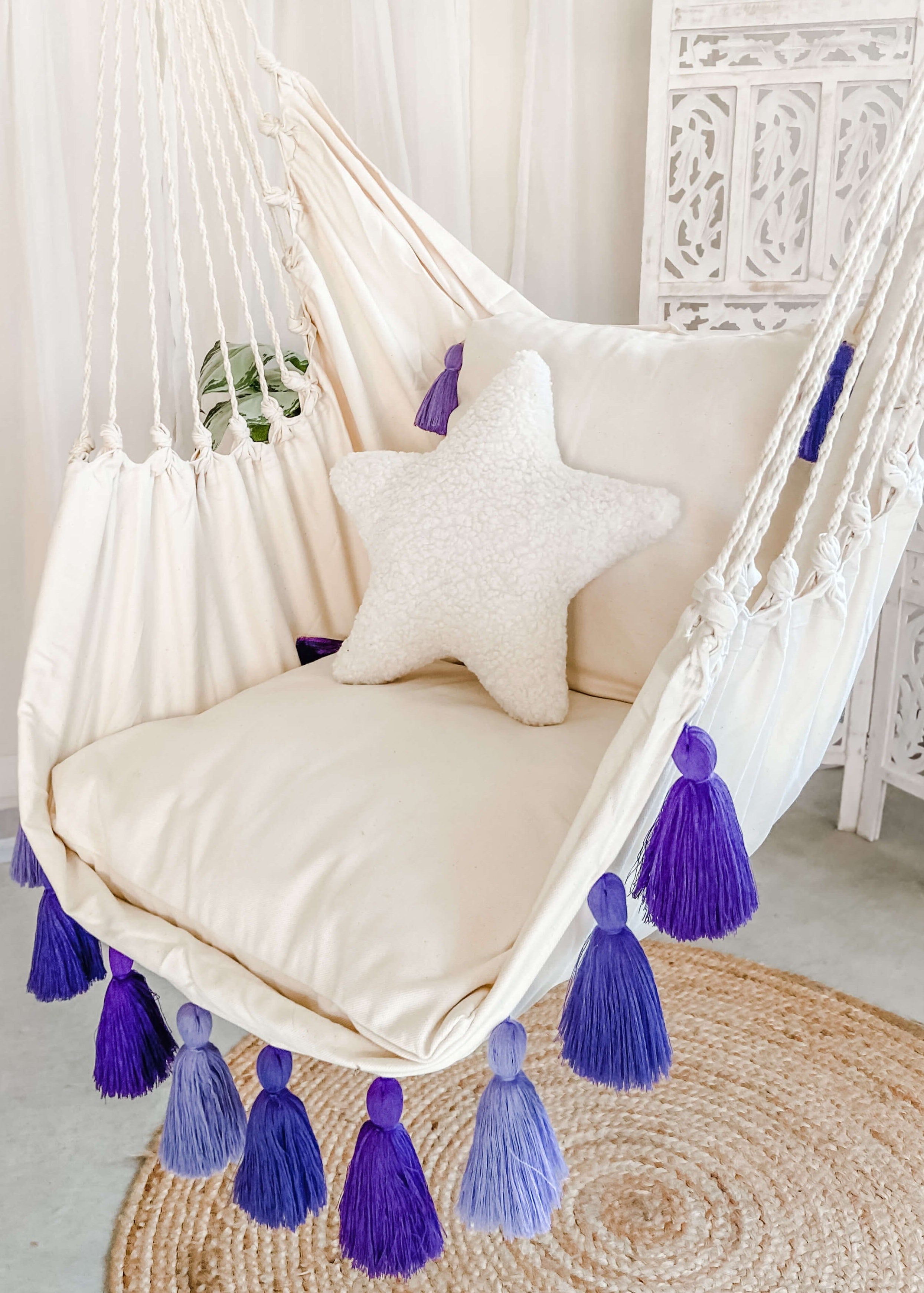 White hammock chair with purple tassels and cushions on a light wooden floor.