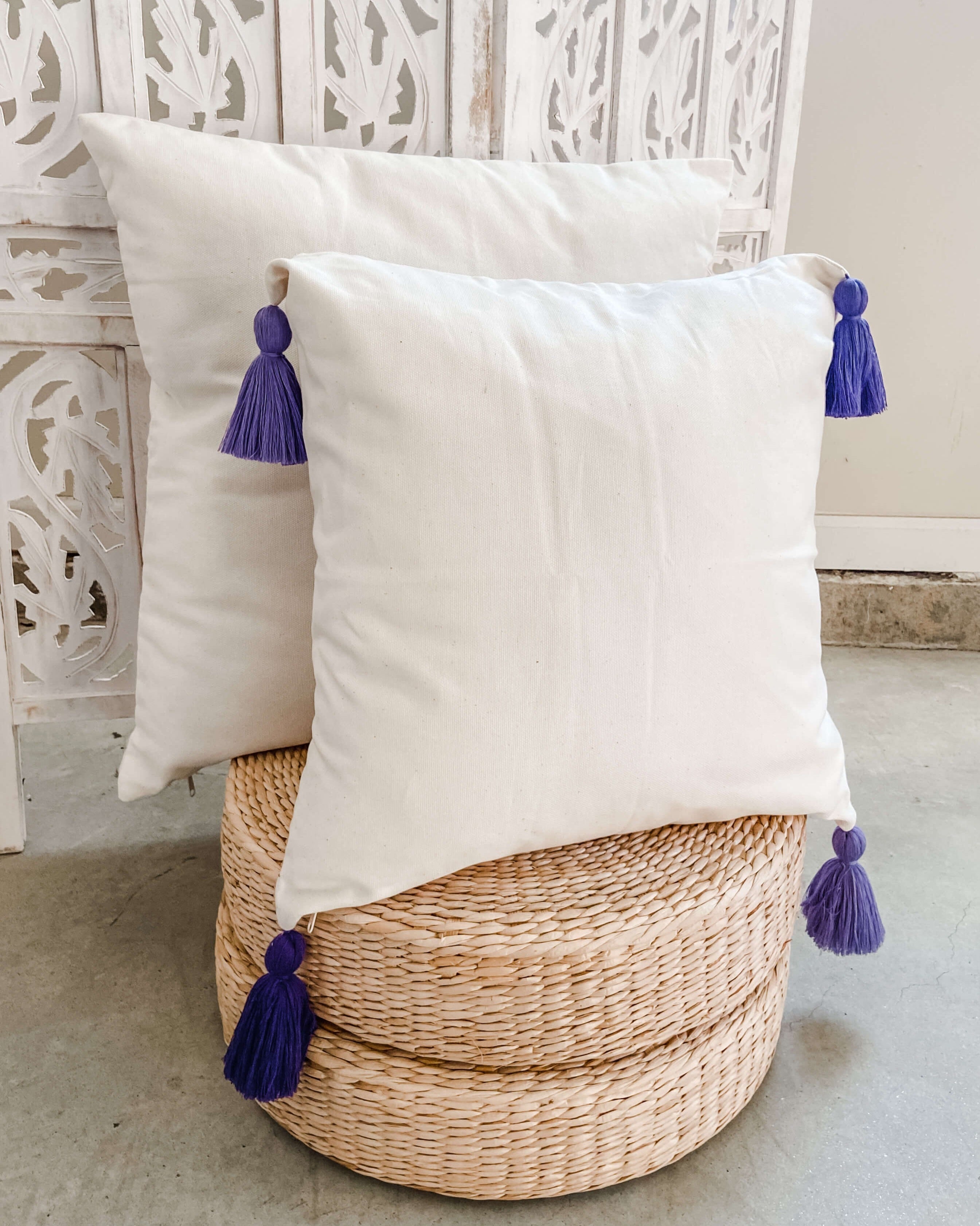 White pillows with purple tassels on a woven stool against a decorative wall.
