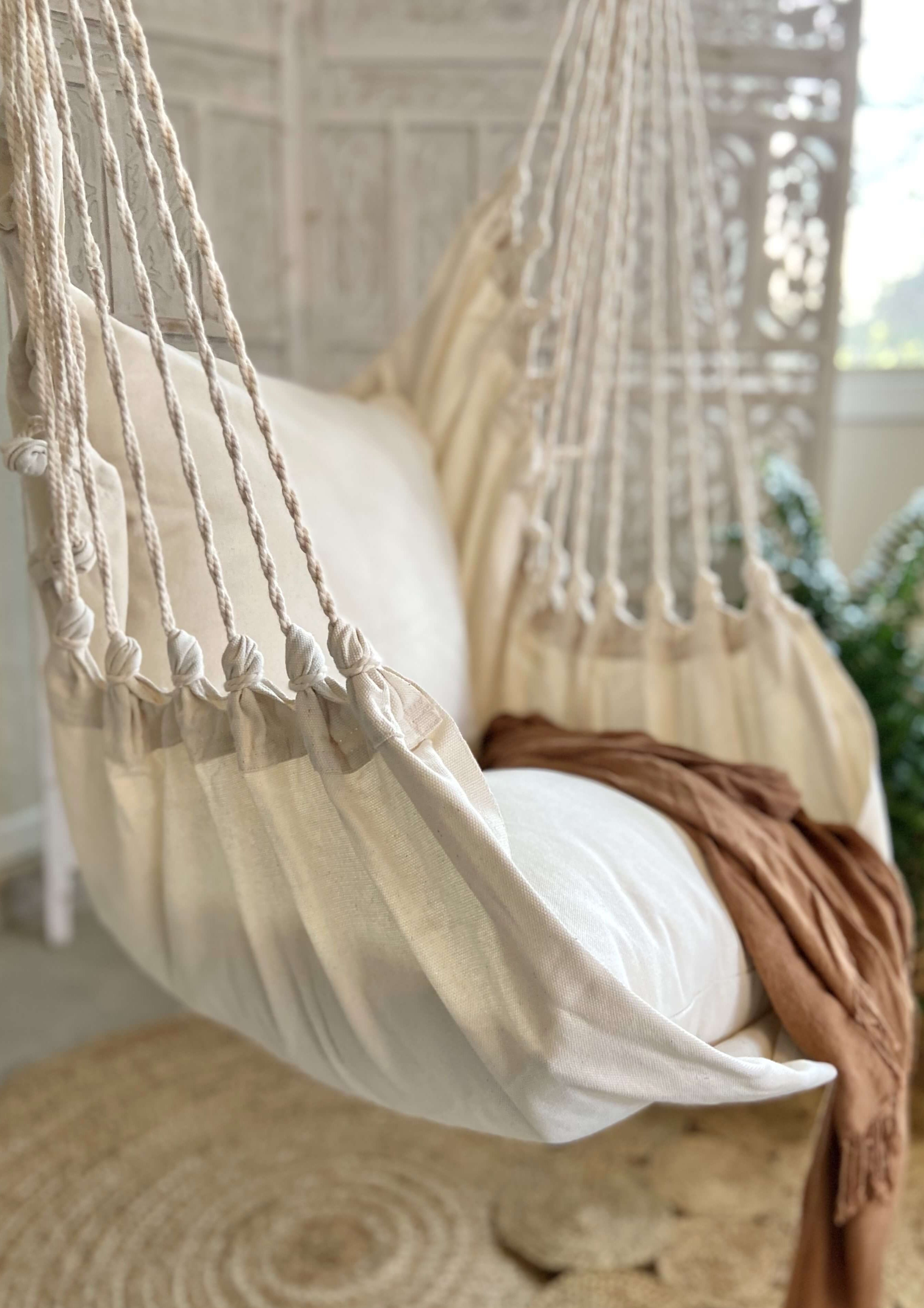 Beige hammock chair with white cushion and brown blanket in a room with plants and window.