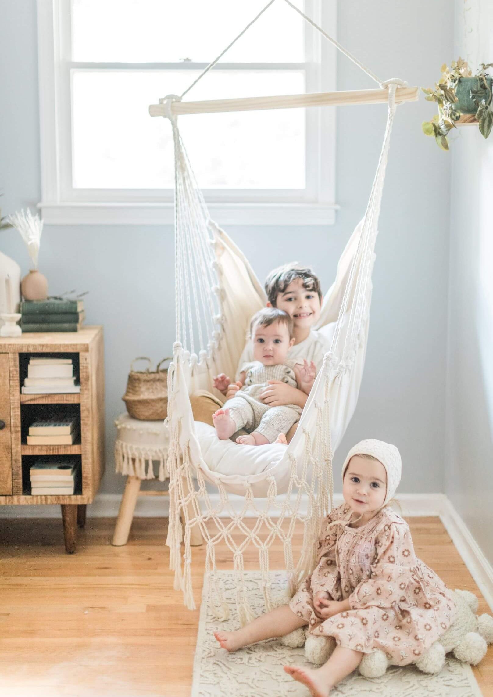 Children sitting in a macrame hanging chair in a bright room with wooden floor and light blue walls.