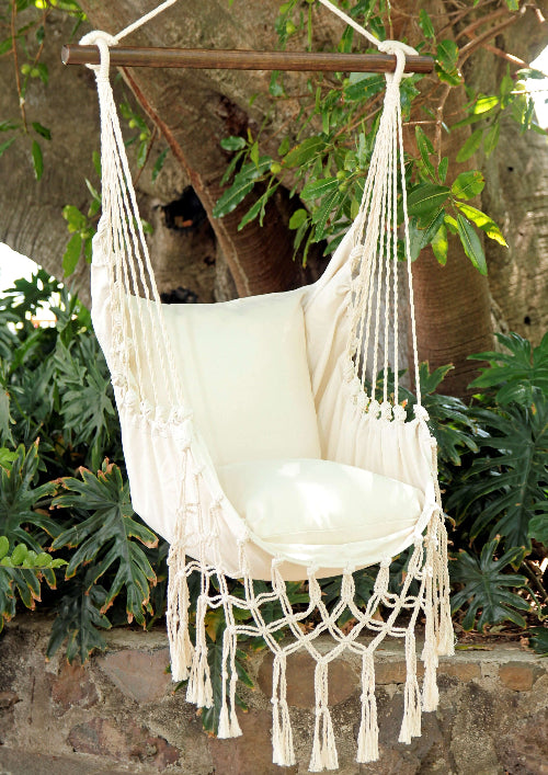 White macrame hammock chair with tassels hanging against a natural background with plants.