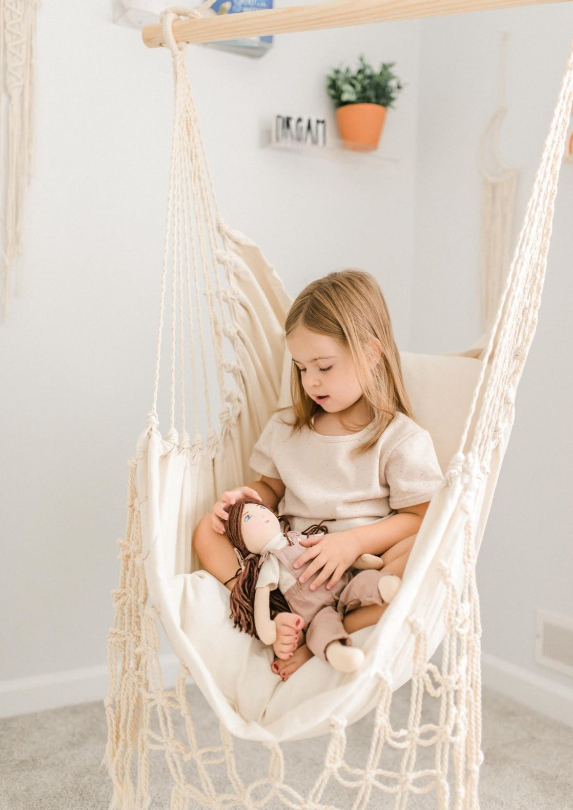 Child sitting in a macrame hammock holding a plush toy, with a neutral background