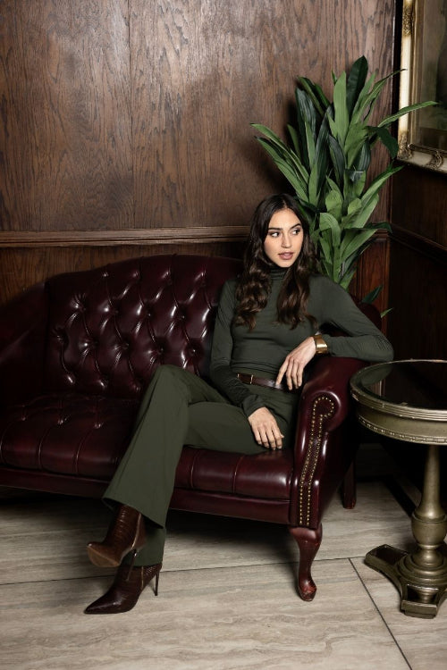 Woman in a green outfit sitting on a leather couch in a room with wooden walls and a plant.