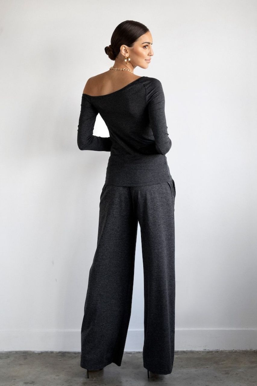 Woman wearing a black off-shoulder top and gray wide-leg pants against a white wall.