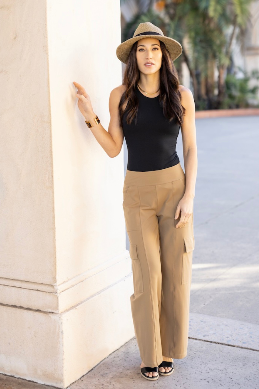 Woman in black top and beige wide-leg pants standing against a wall outdoors.