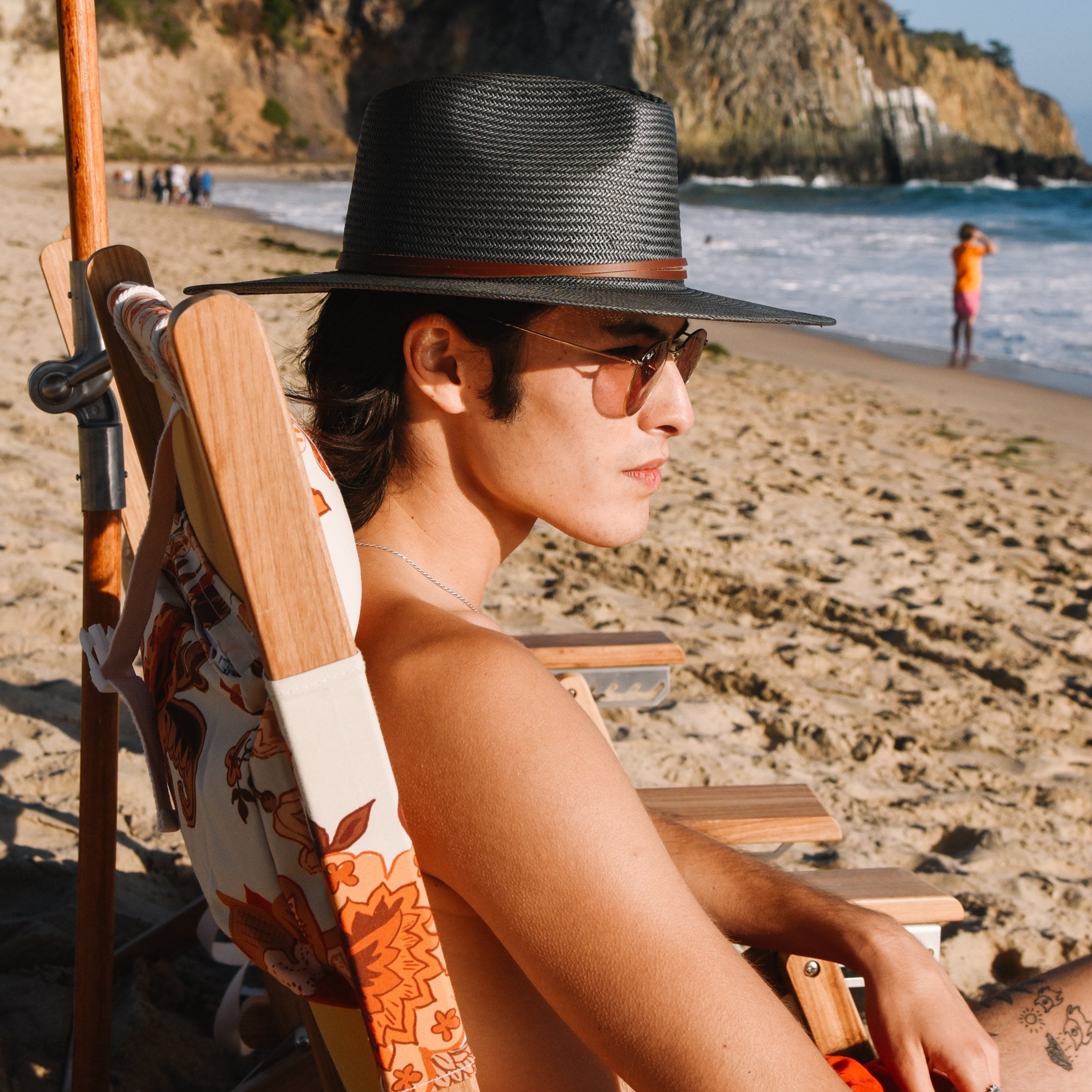 Person sitting on a beach chair by the ocean, wearing a black hat and sunglasses.