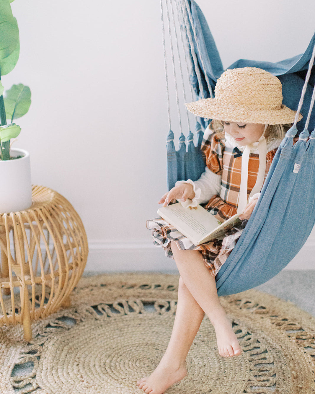 Child reading a book in a blue hammock with a plant and side table in the background