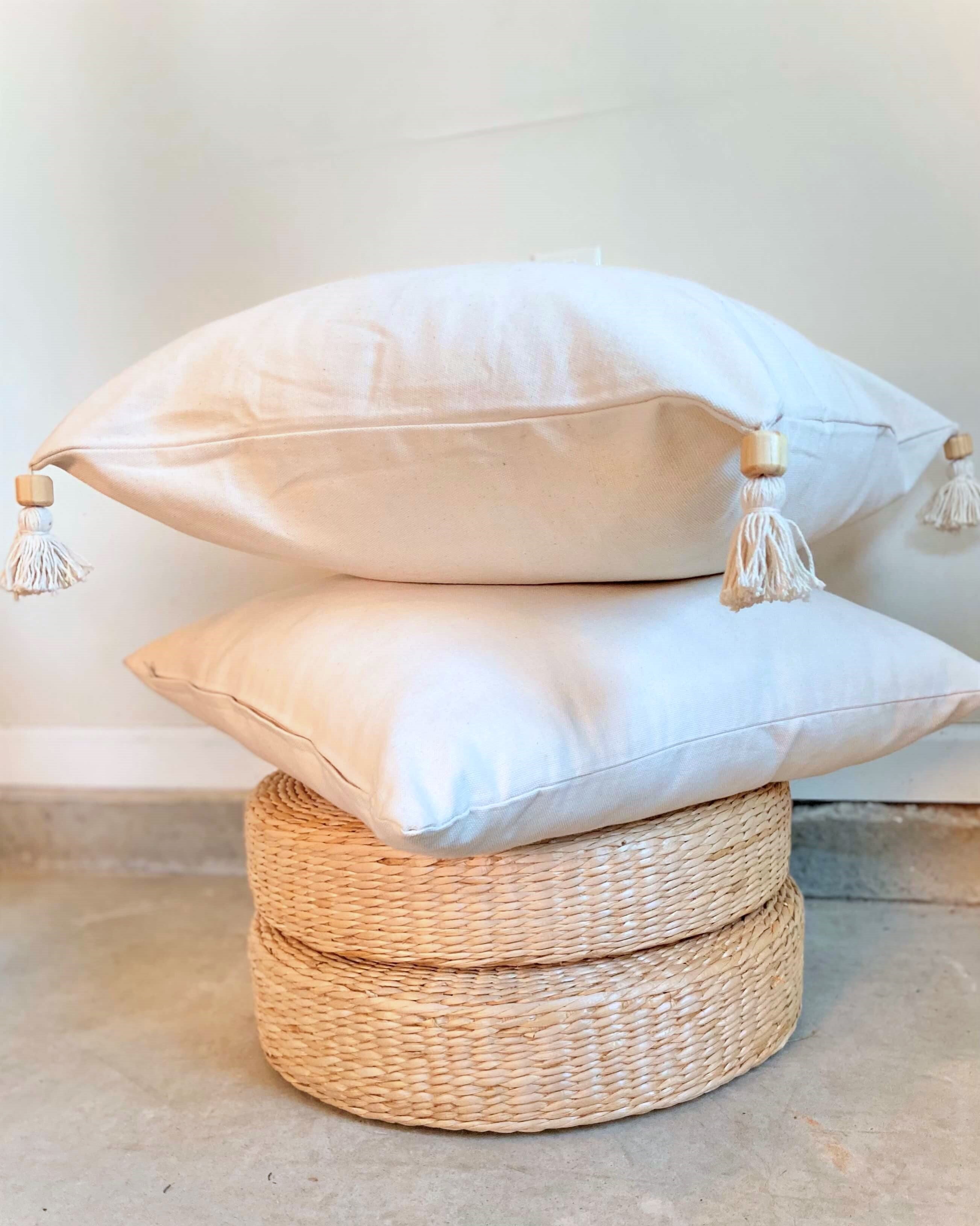 Two white pillows with tassels on a woven stool against a light background