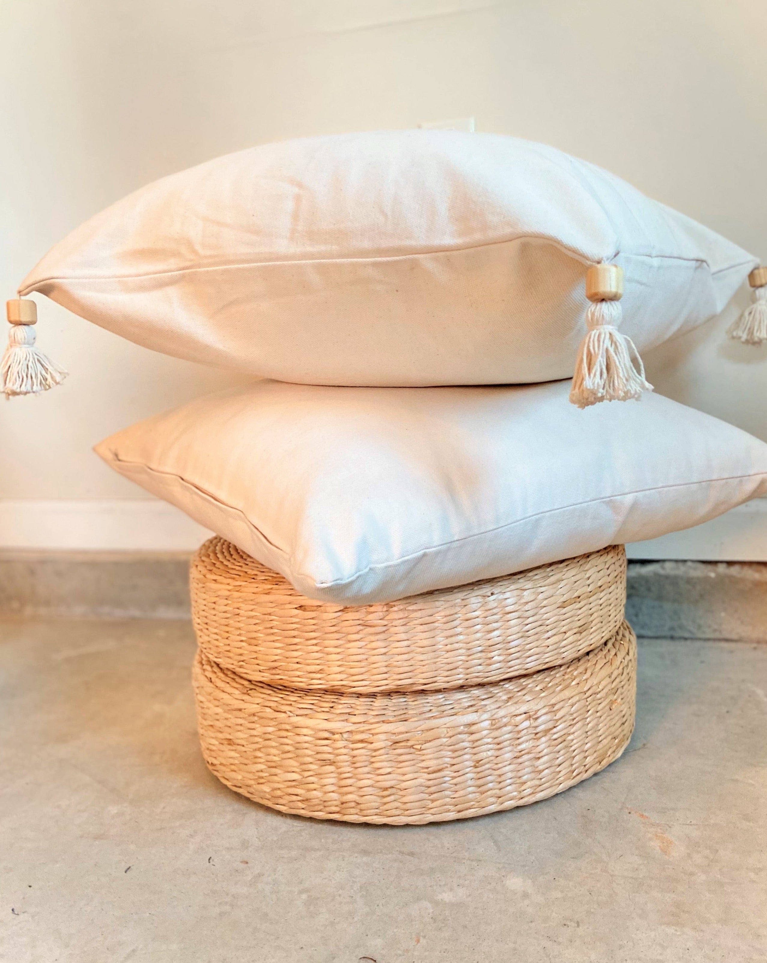 Stack of decorative pillows with tassels on a woven ottoman against a neutral background