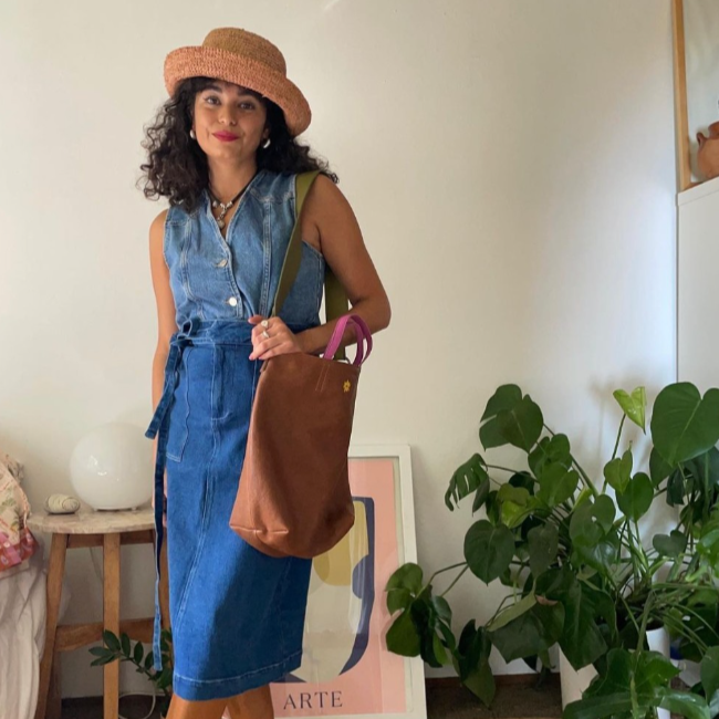 Woman in a blue dress and straw hat holding a brown bag indoors with plants and decor.