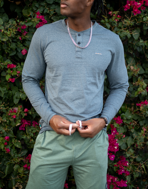 Person wearing a blue long-sleeve shirt and green pants standing in front of pink flowers.