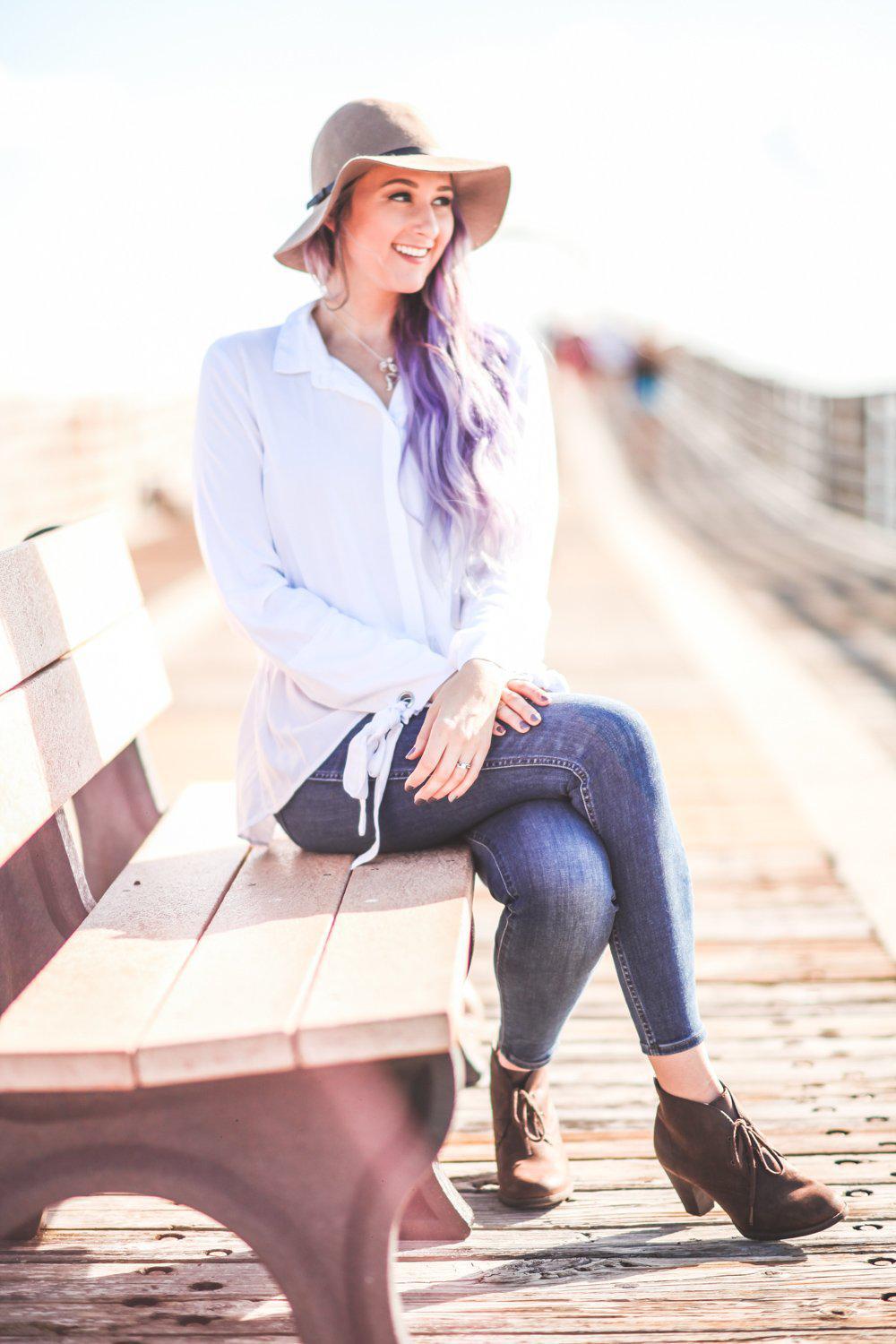 Woman sitting on a wooden bench on a boardwalk wearing a white shirt, blue jeans, and a hat.