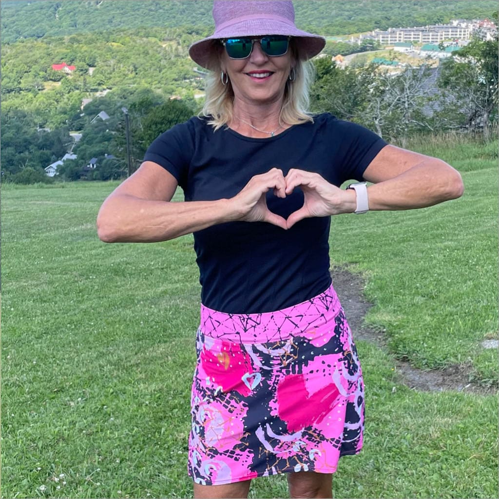 Woman in black t-shirt and colorful skirt making a heart shape with her hands outdoors.