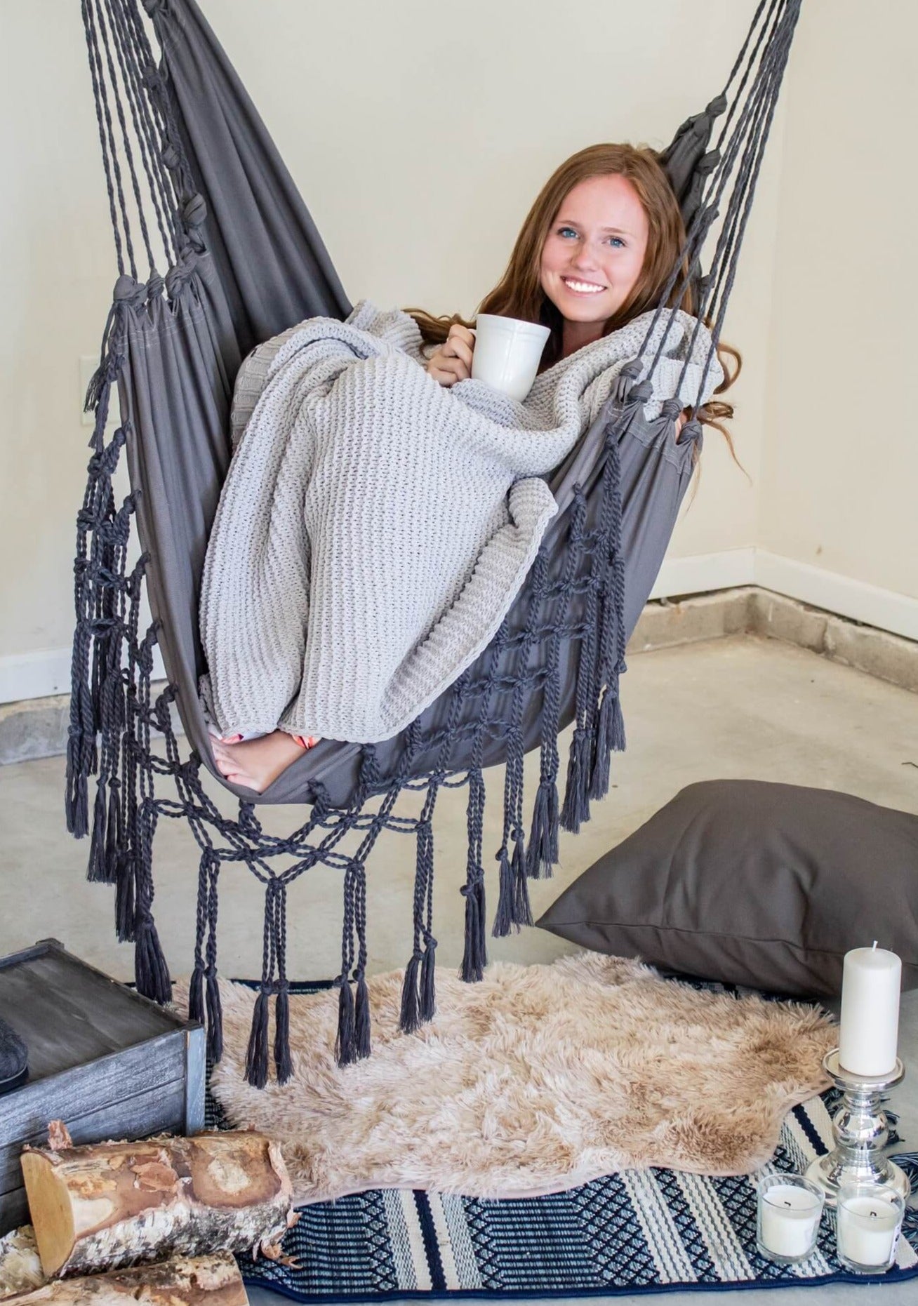 Woman sitting in a gray hammock chair holding a mug, surrounded by cozy decor.