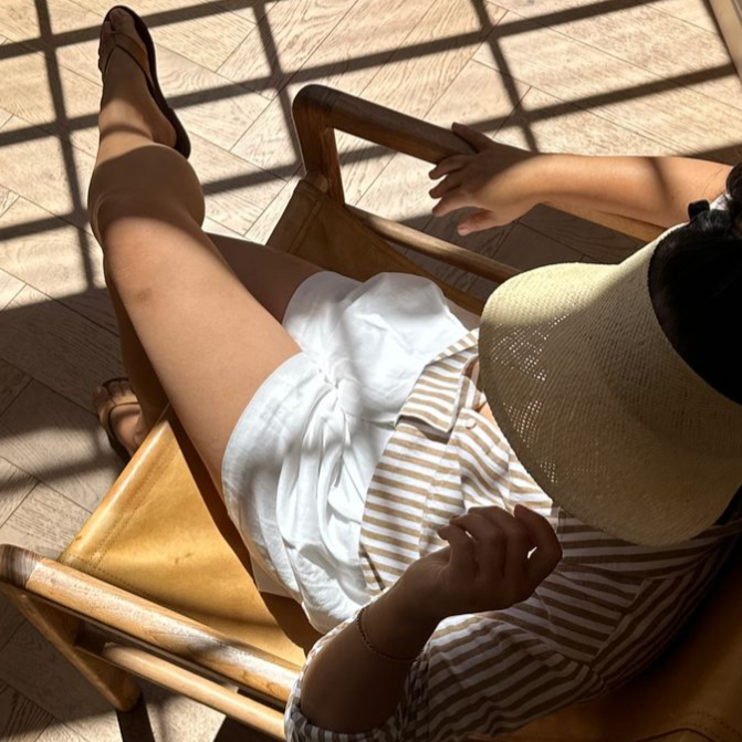 Person sitting on a chair with legs crossed, wearing white pants and a striped shirt, in a sunlit room.