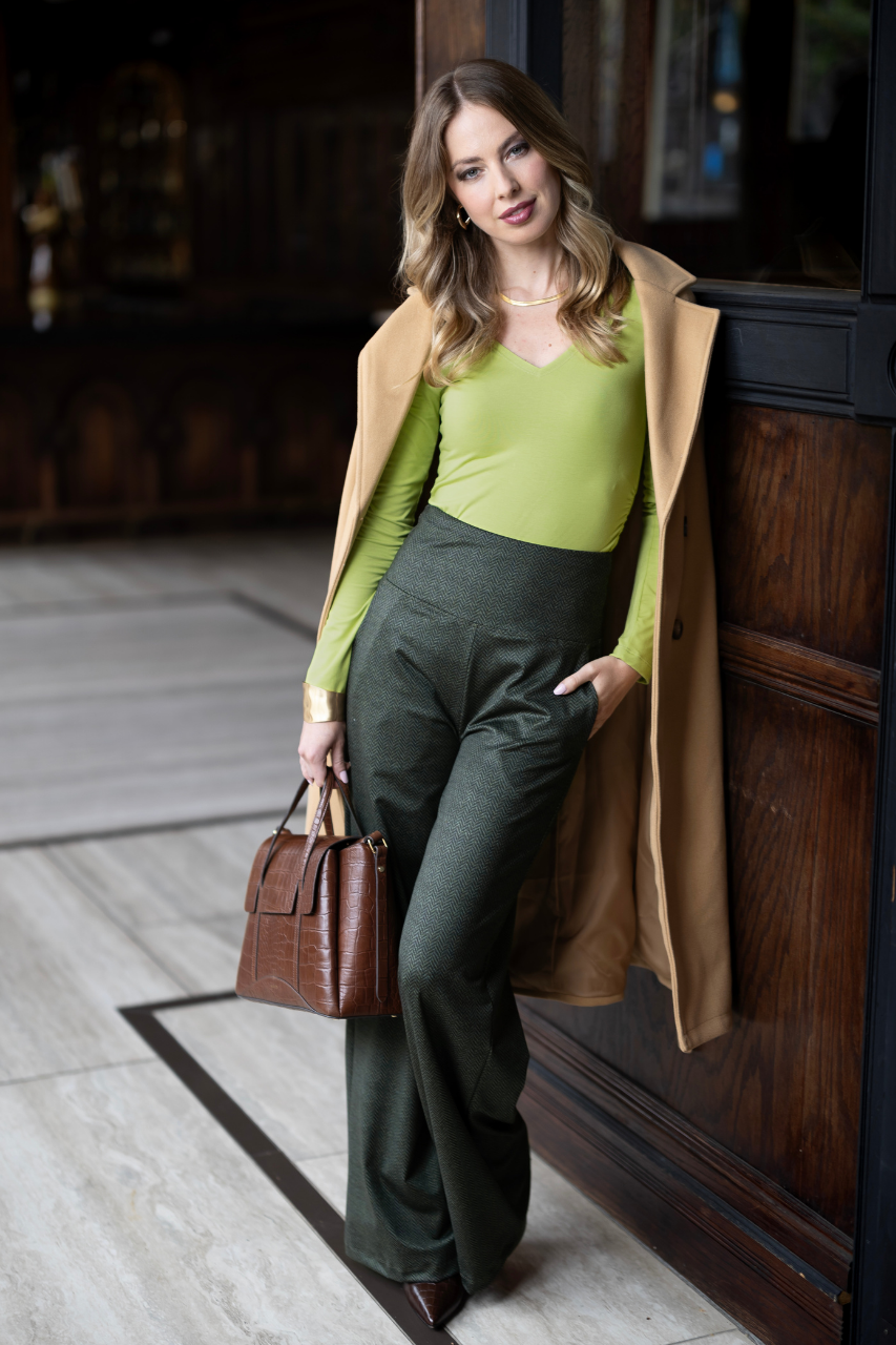 Woman in green top and dark pants with a tan coat and brown handbag standing against a wooden door.