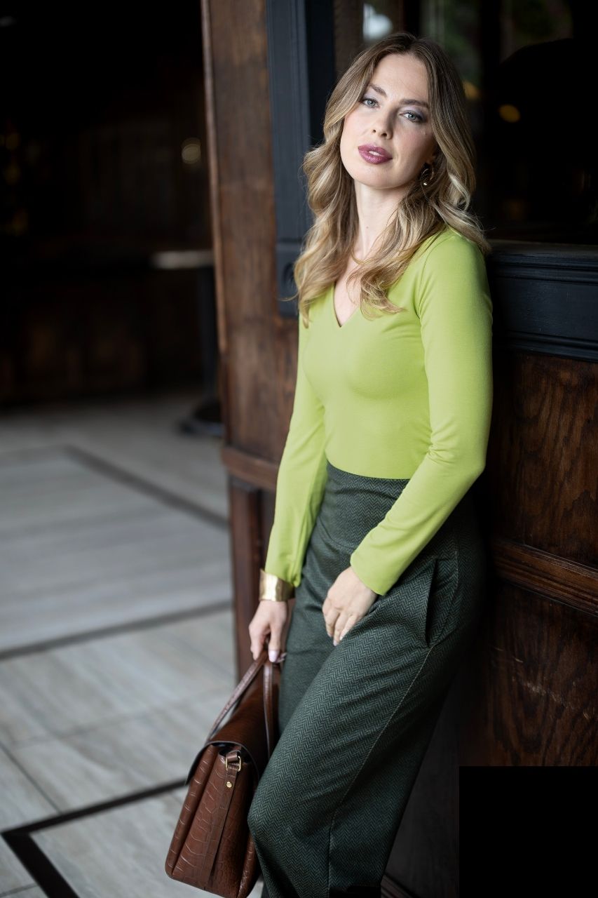 Woman in a green outfit holding a brown bag against a wooden wall.