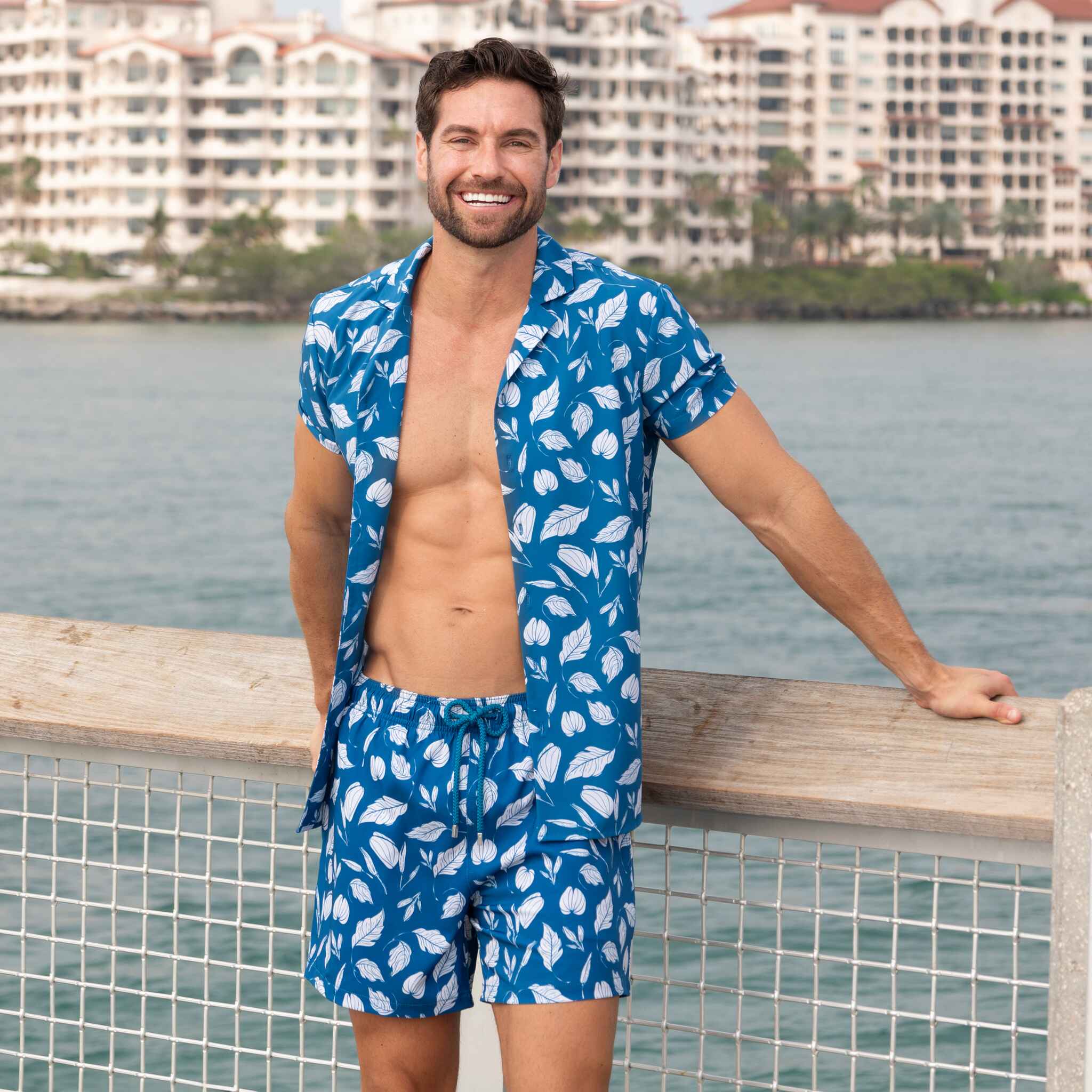 Man wearing a blue patterned shirt and shorts by a waterfront with buildings in the background