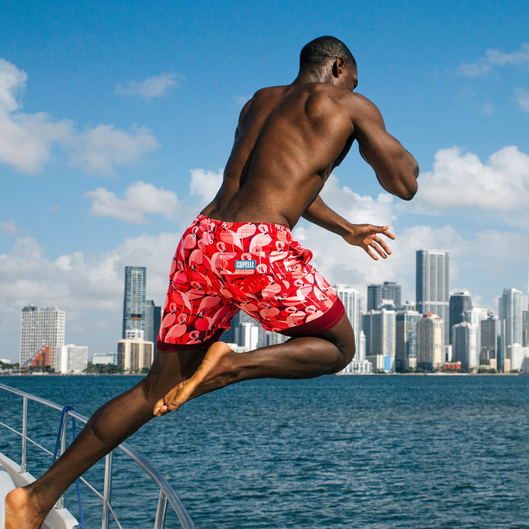 Man in red swim shorts jumping off a boat with a city skyline in the background