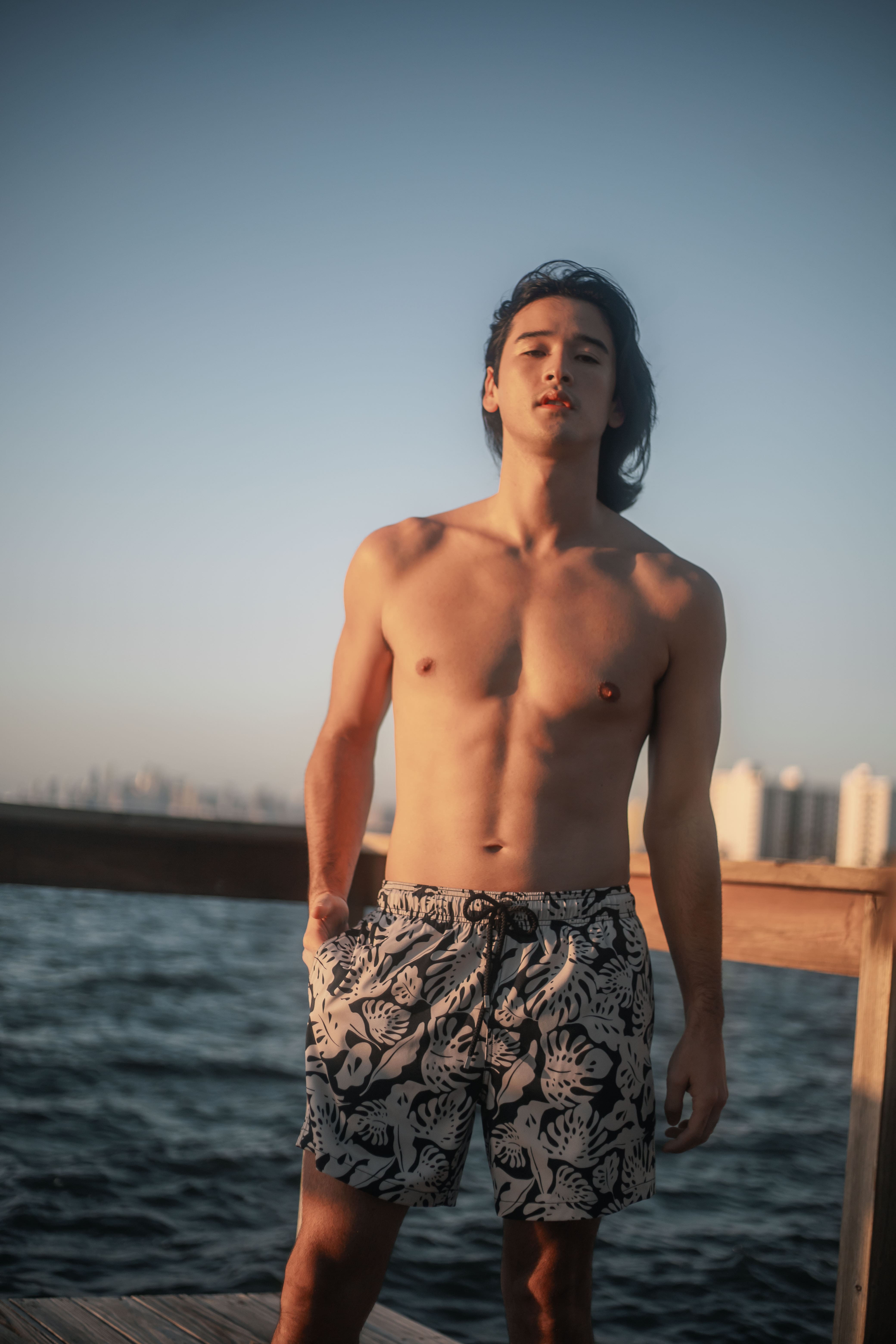 Man standing by a waterfront wearing patterned shorts with a city skyline in the background.