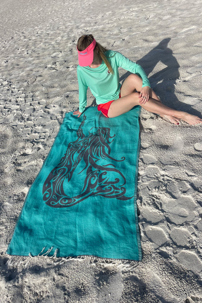 Person sitting on a teal towel with a dragon design on a sandy beach.