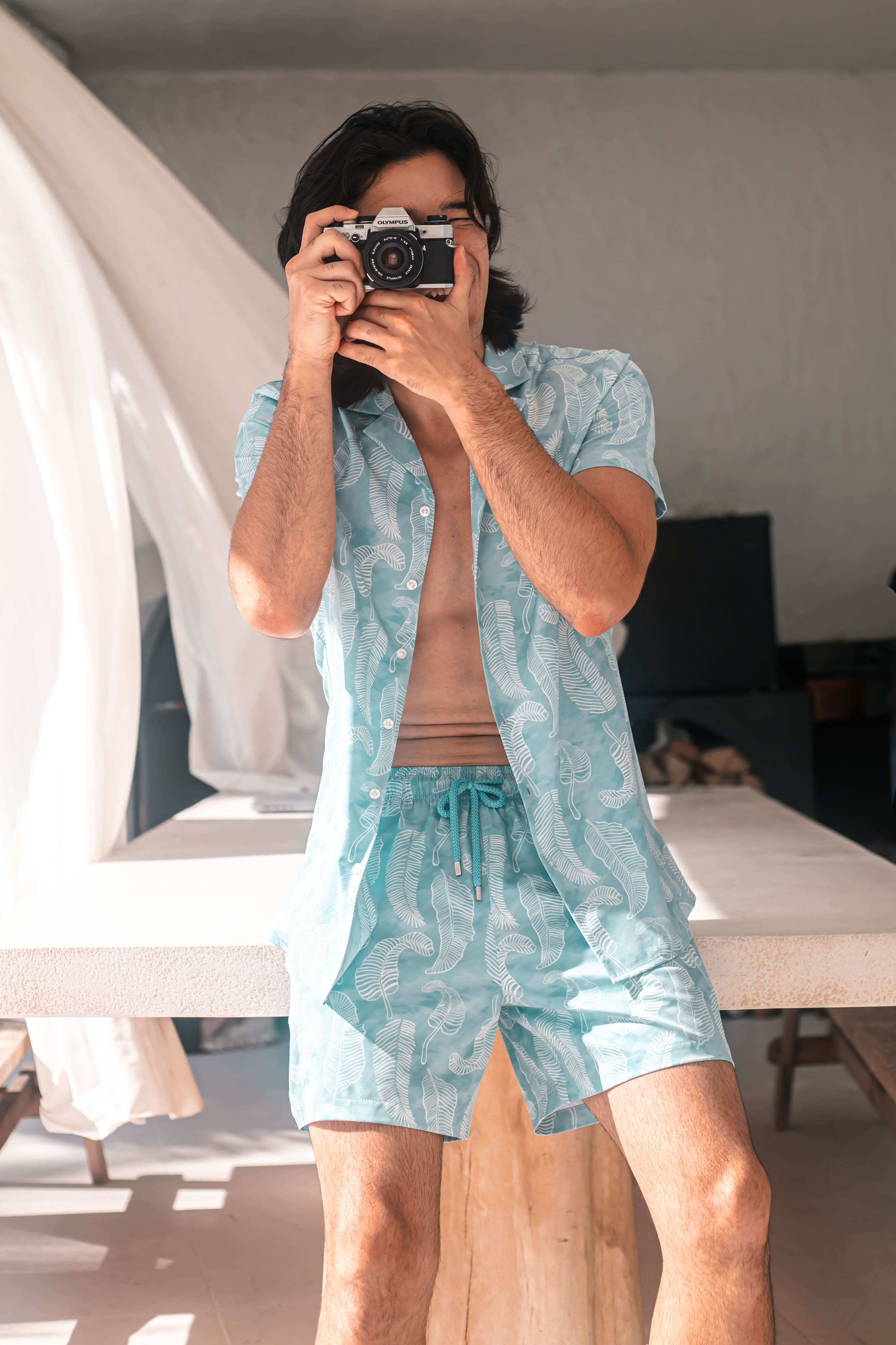 Person taking a mirror selfie wearing a light blue patterned shirt and shorts.