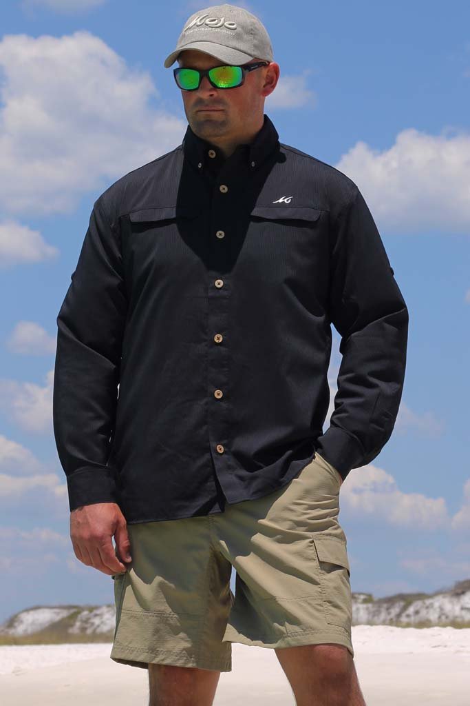 Man wearing a black jacket, beige cap, and sunglasses on a beach.