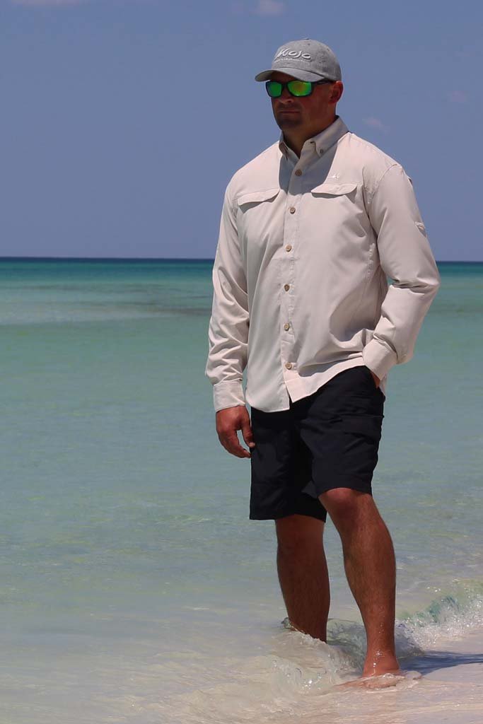 Man standing in shallow water at the beach wearing a light-colored shirt, dark shorts, and sunglasses.