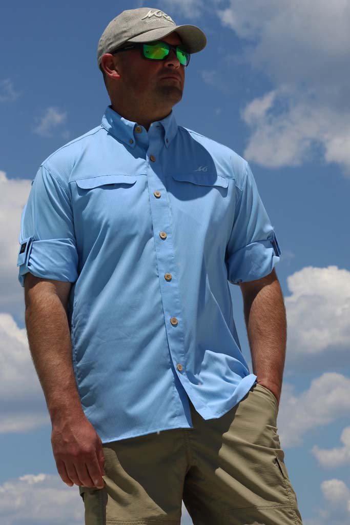 Man wearing a light blue shirt, beige cap, and sunglasses against a blue sky with clouds.
