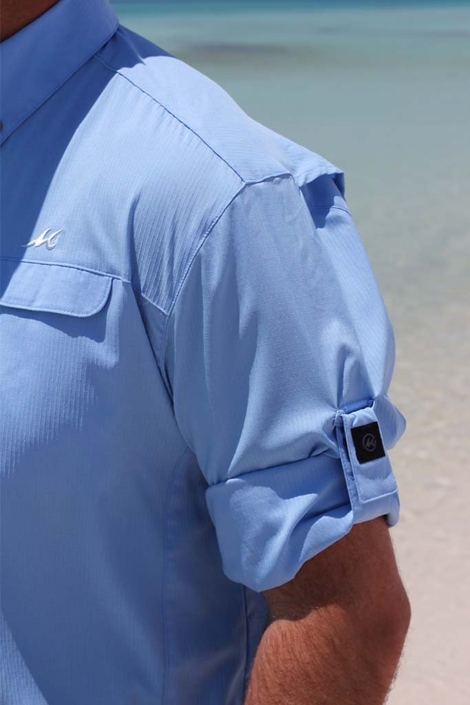 Close-up of a person wearing a light blue shirt with rolled-up sleeves on a beach.
