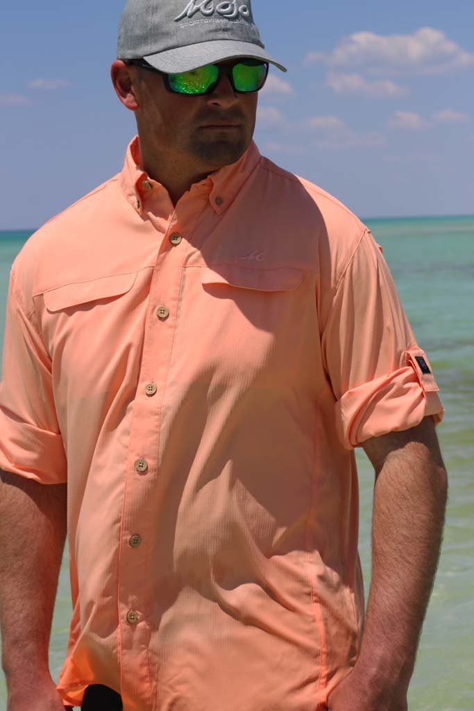 Man wearing a peach shirt, gray cap, and green sunglasses by the beach.