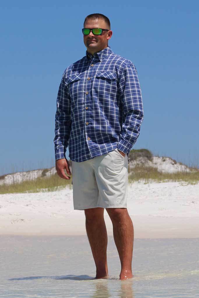Man wearing a blue plaid shirt and light shorts standing on a beach.
